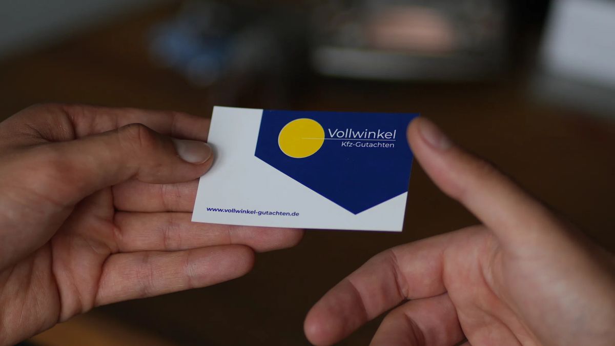 Zwei Hände übergeben eine Visitenkarte mit dem Logo von Vollwinkel Kfz-Gutachten, dahinter unscharf ein Schreibtisch.