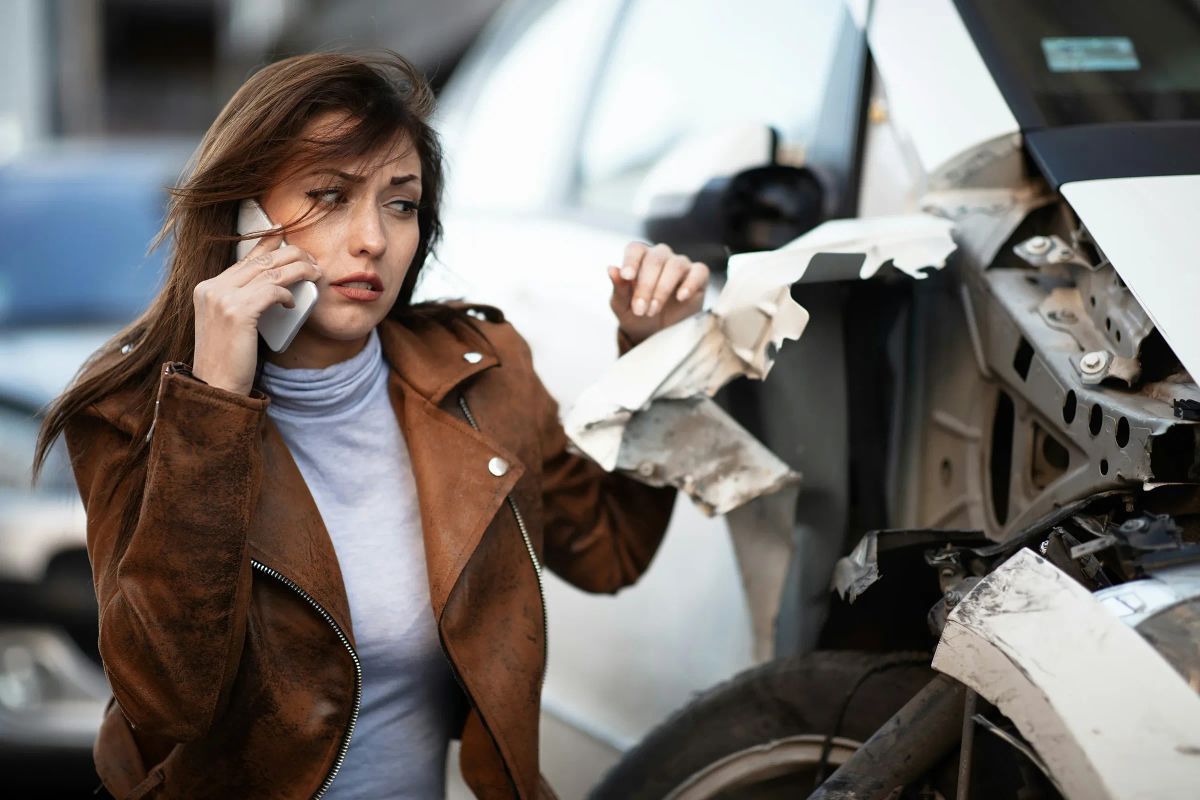 Junge Frau telefoniert an der Straße und blickt auf ein weißes Auto mit aufgerissenem Frontblech und abgerissenem Stoßfänger.