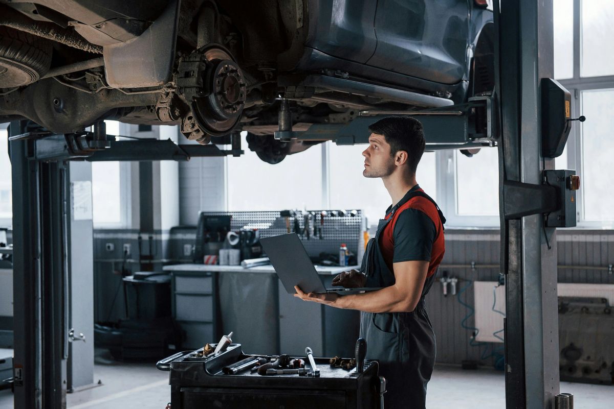 Mechaniker steht mit aufgeklapptem Laptop unter einem Auto auf der Hebebühne und sieht nach oben zum Unterboden.