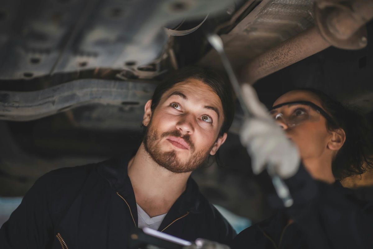 Zwei Mechaniker in Arbeitskleidung prüfen mit Werkzeug den Unterboden eines Autos auf der Hebebühne.