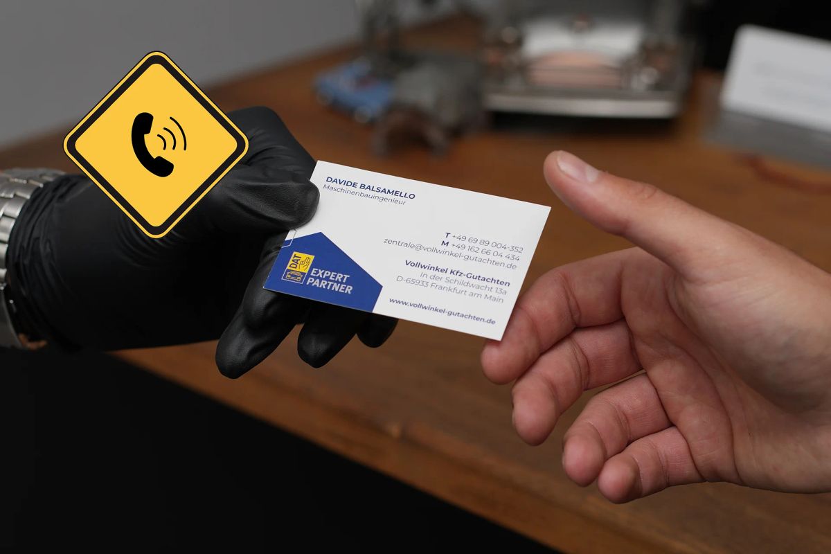 Eine Hand im schwarzen Arbeitshandschuh reicht eine Visitenkarte von Vollwinkel weiter, daneben ein gelbes Schild mit Telefonsymbol