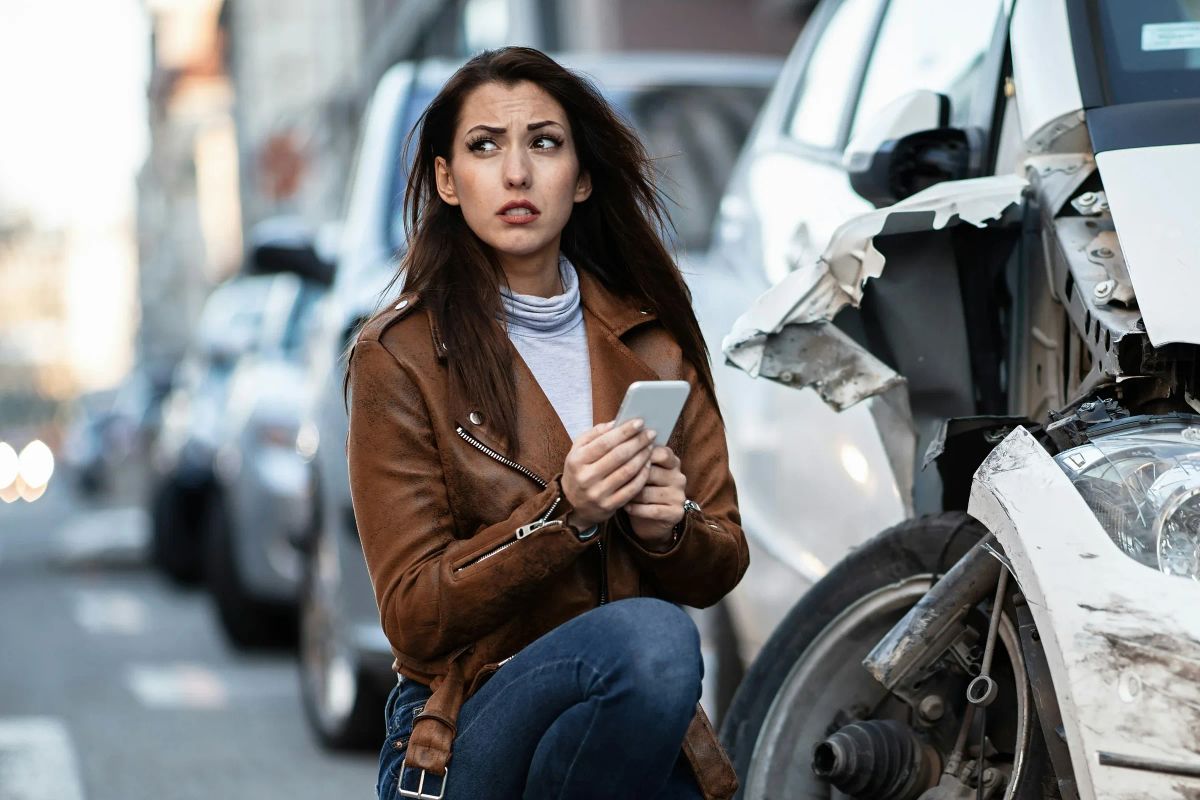 Eine Frau hockt mit dem Smartphone am Straßenrand neben einem weißen Auto, dessen Front aufgerissen und dessen Vorderrad abgeknickt ist.