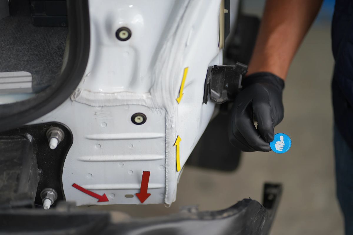Gelbe und rote Pfeile markieren gestauchtes Blech hinter dem abgebauten Stoßfänger eines weißen Transporters, daneben eine Hand im schwarzen Handschuh.