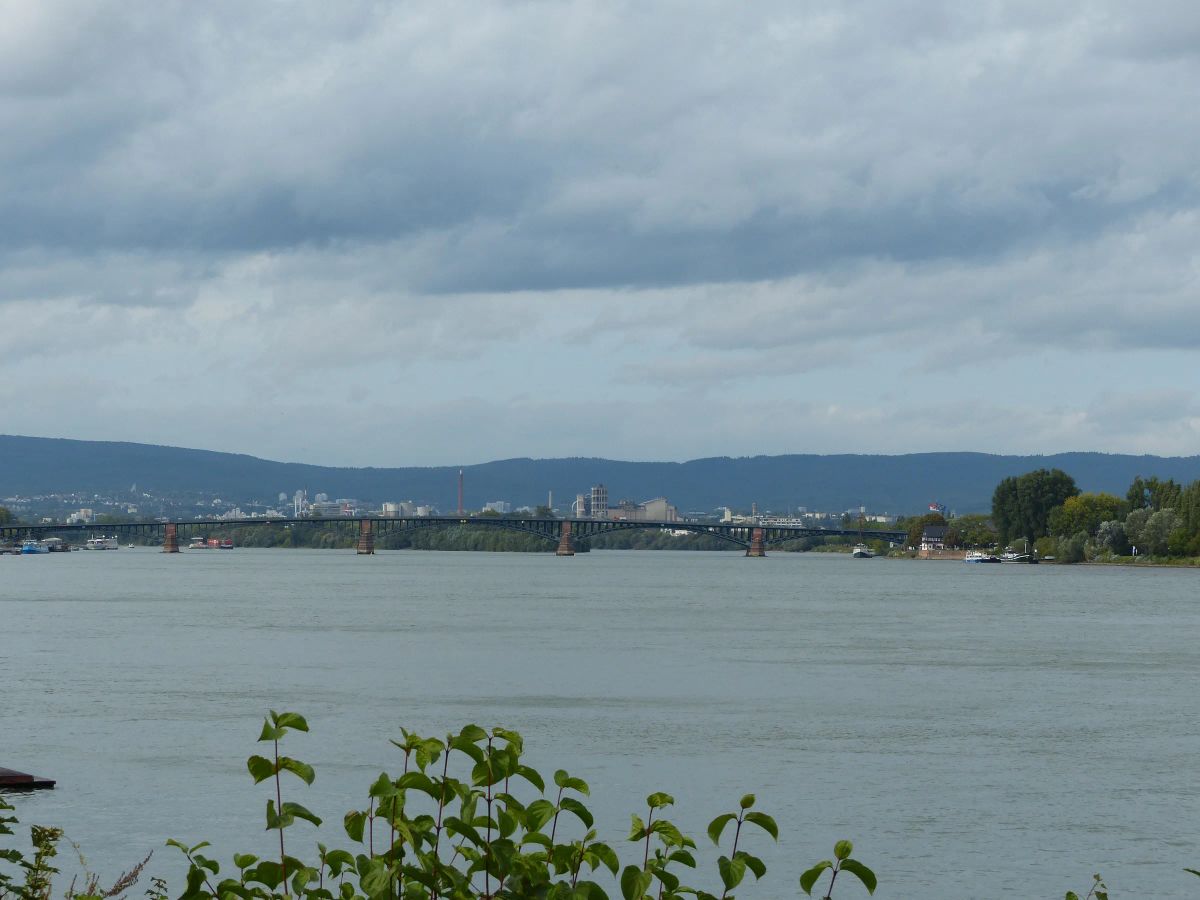 Die Theodor-Heuss-Brücke spannt sich in flachen Bögen über den Rhein, dahinter Gebäude am anderen Ufer und die Höhen des Taunus unter Wolken.