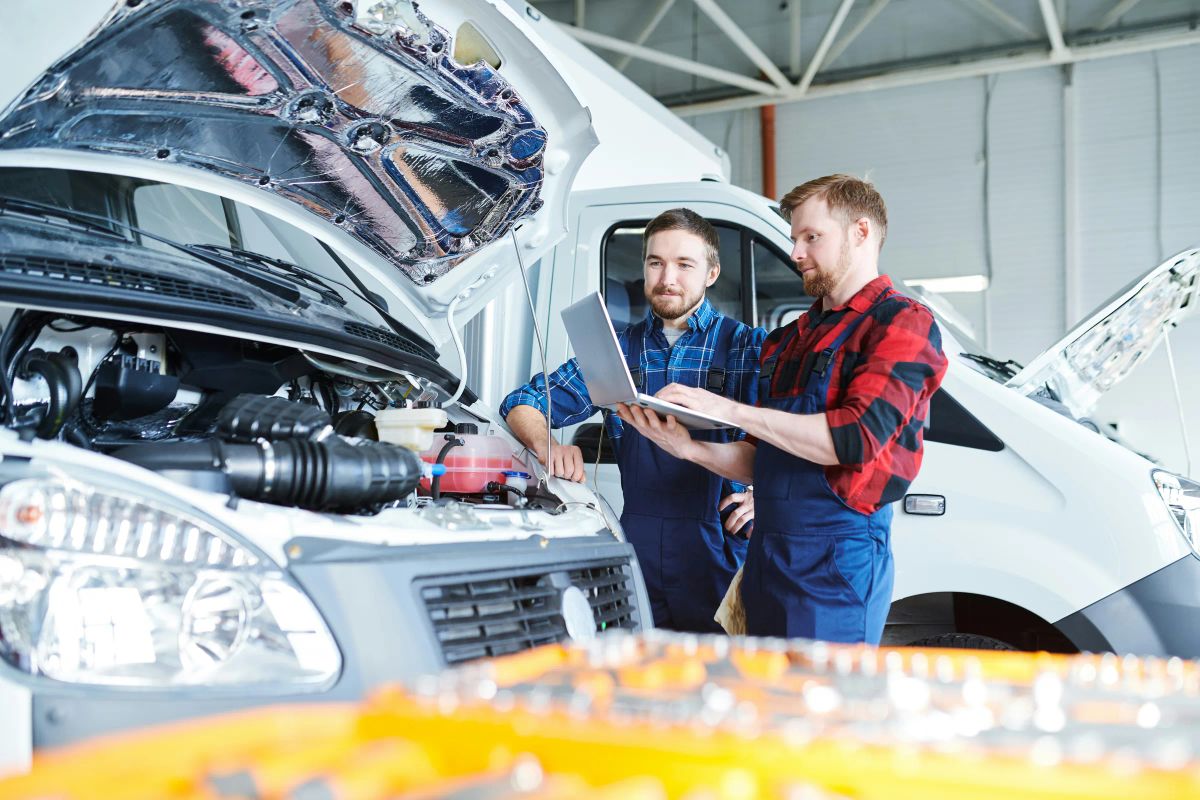 Zwei Mechaniker in blauen Latzhosen stehen vor der geöffneten Motorhaube eines weißen Transporters und schauen gemeinsam auf einen Laptop.