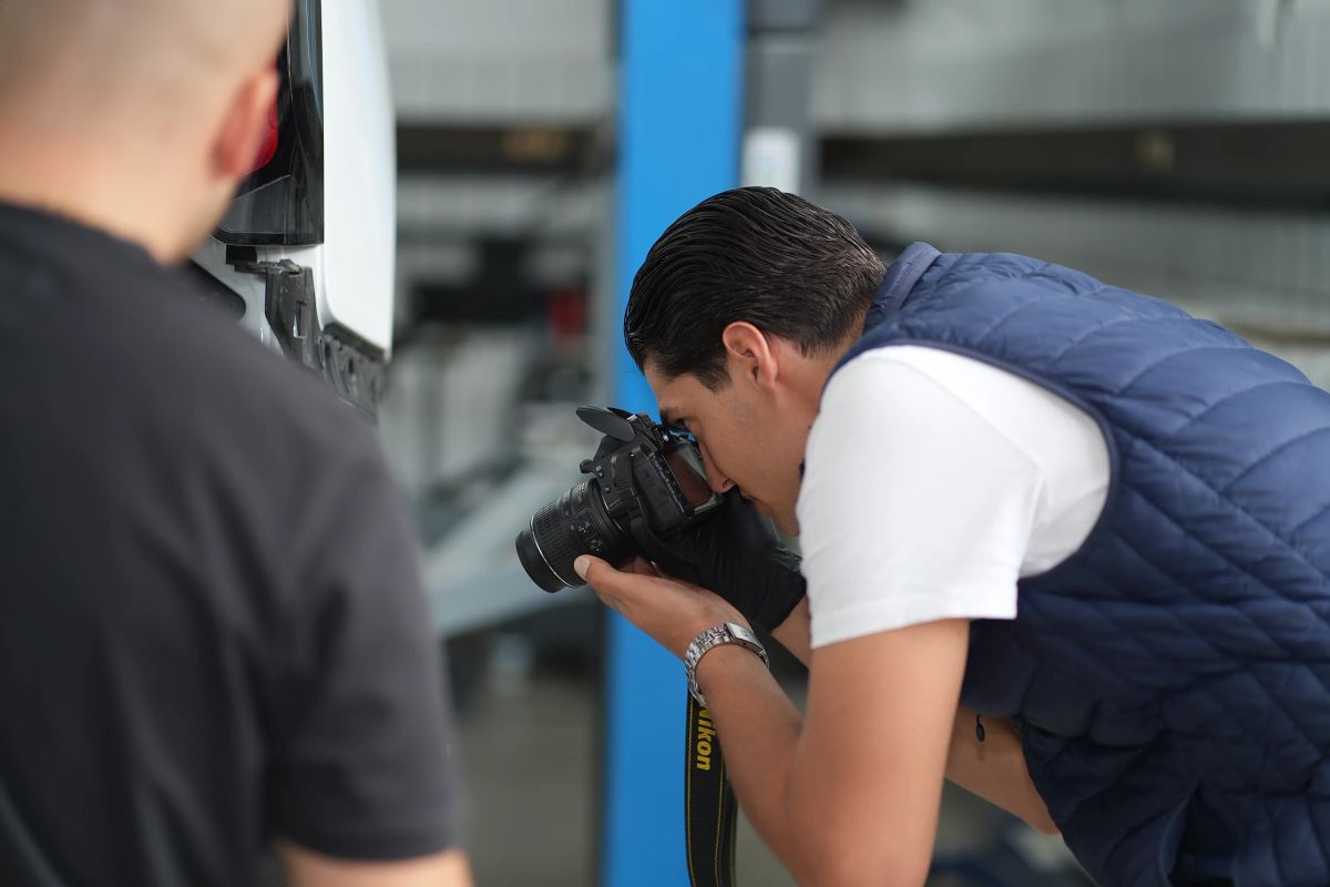 Sachverständiger fotografiert mit einer Spiegelreflexkamera den Schaden an einem weißen Kastenwagen, der Kunde schaut über die Schulter zu.